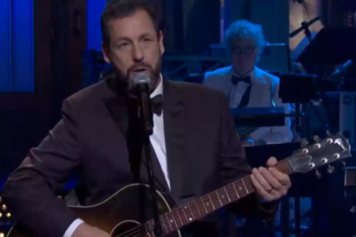 Un hombre con traje oscuro toca la guitarra y canta frente a un micrófono en un escenario iluminado, mientras un músico de fondo toca el teclado.