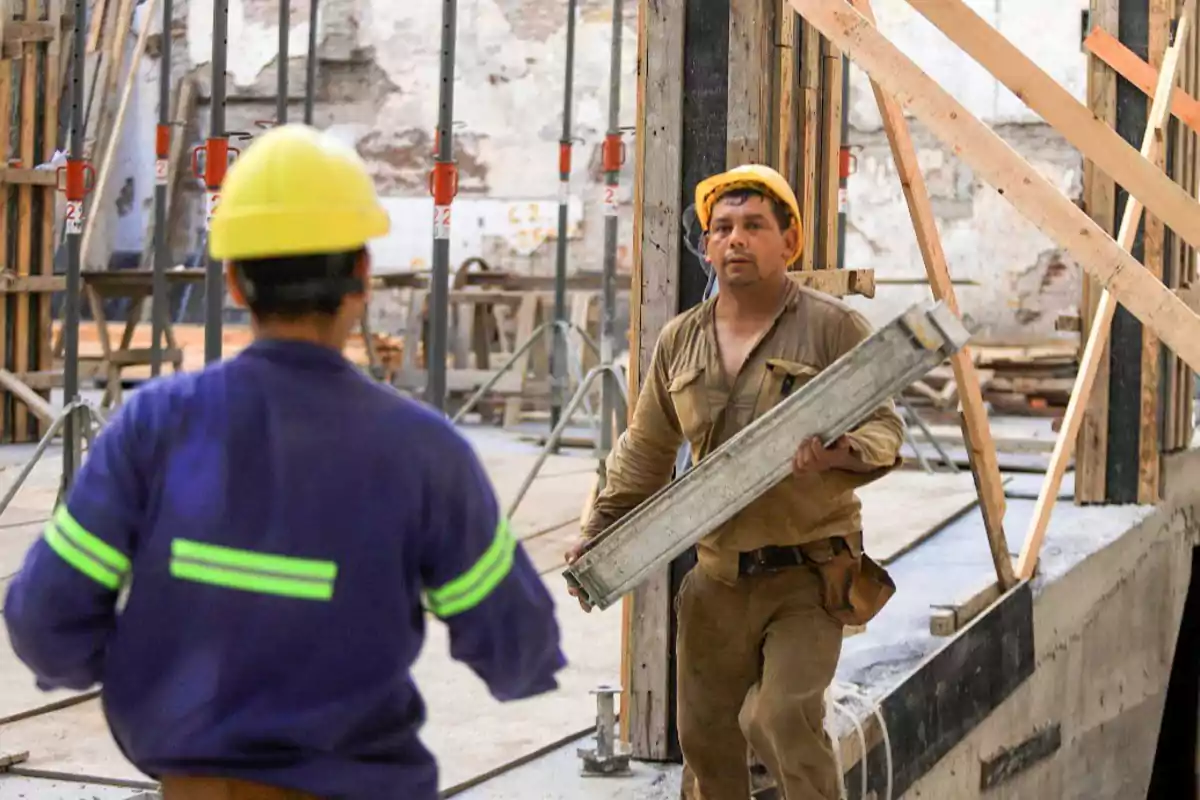 Dos trabajadores de la construcción con cascos amarillos en un sitio de obra, uno de ellos lleva una viga metálica.