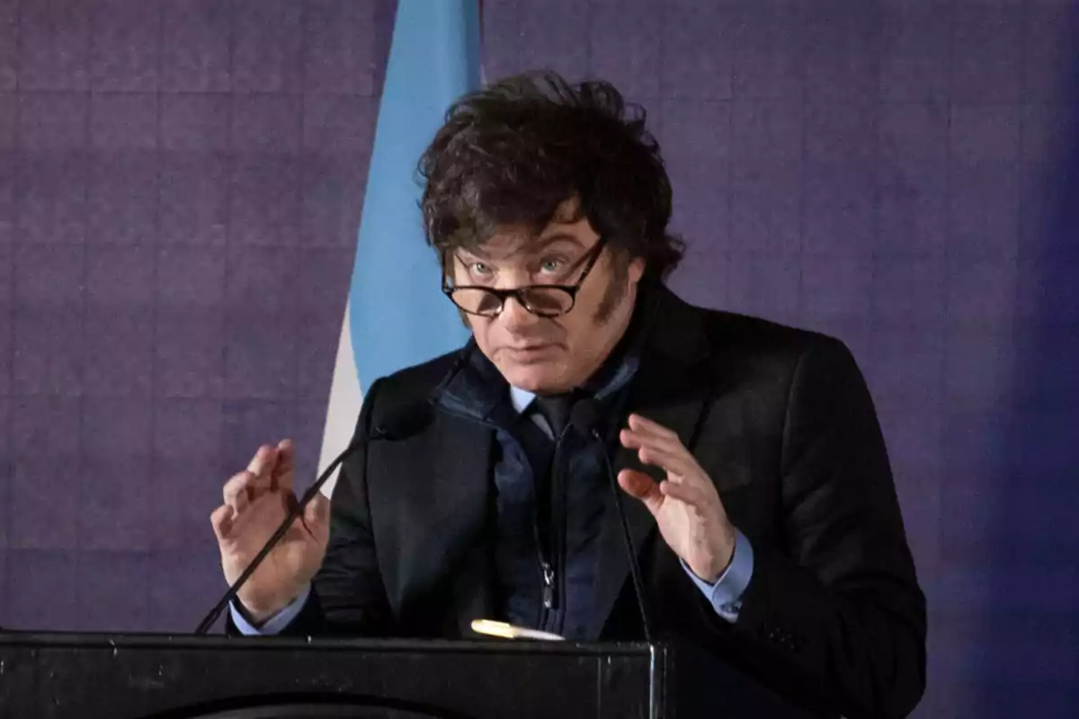 Un hombre con gafas hablando en un podio con una bandera azul y blanca de fondo.