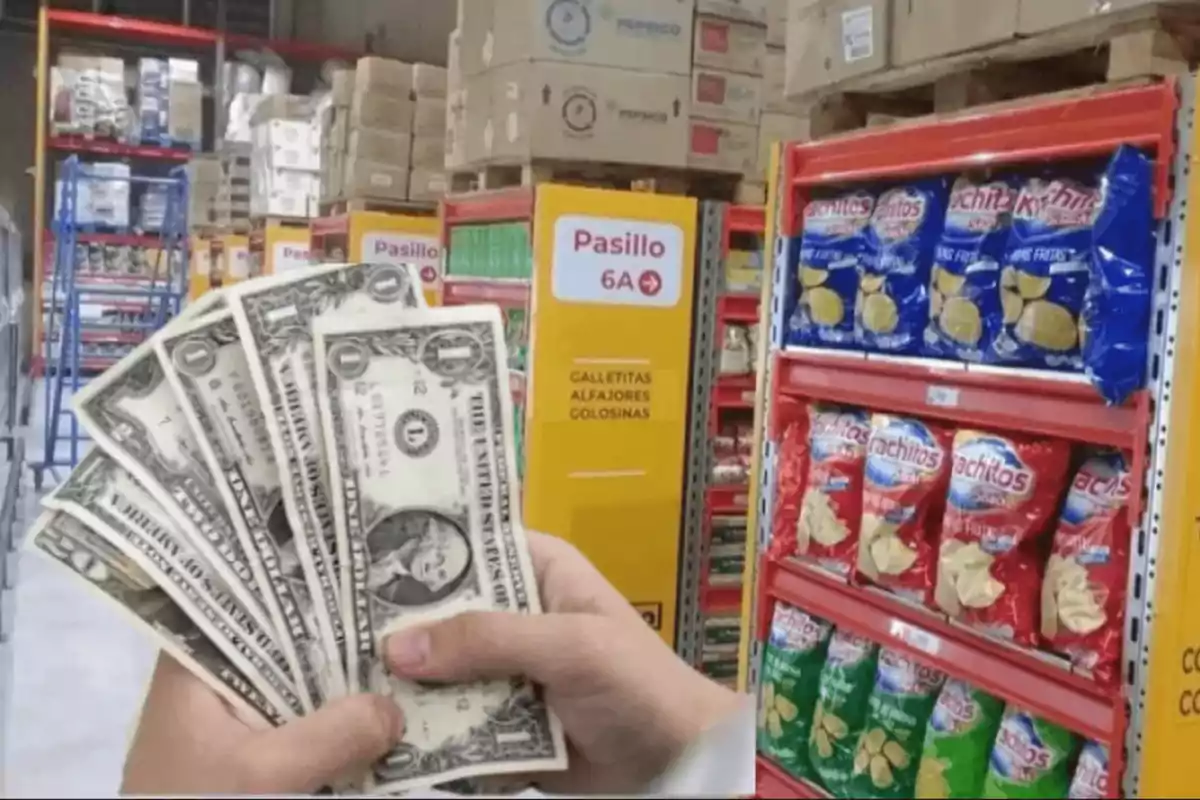 Una mano sosteniendo billetes de un dólar en un pasillo de supermercado con estantes llenos de productos y un letrero que indica "Pasillo 6A" con galletitas, alfajores y golosinas.