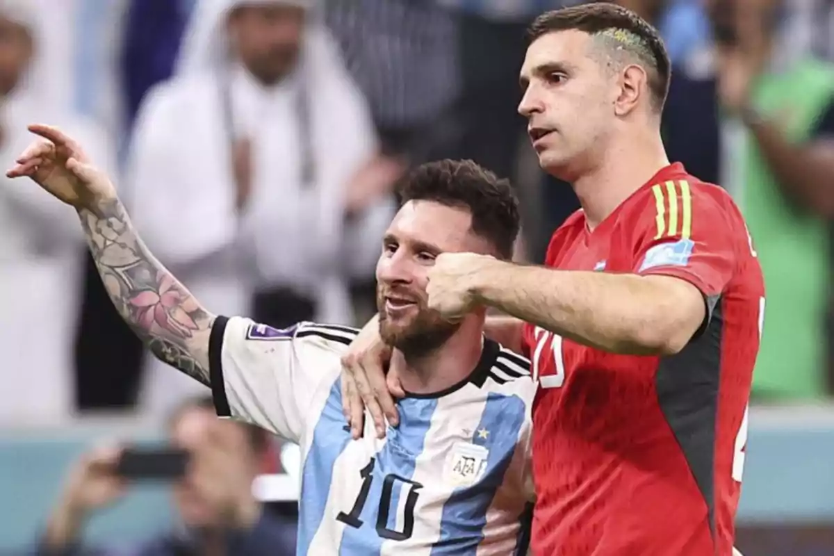 Dos jugadores de fútbol celebran juntos en el campo, uno con uniforme blanco y celeste y el otro con uniforme rojo.