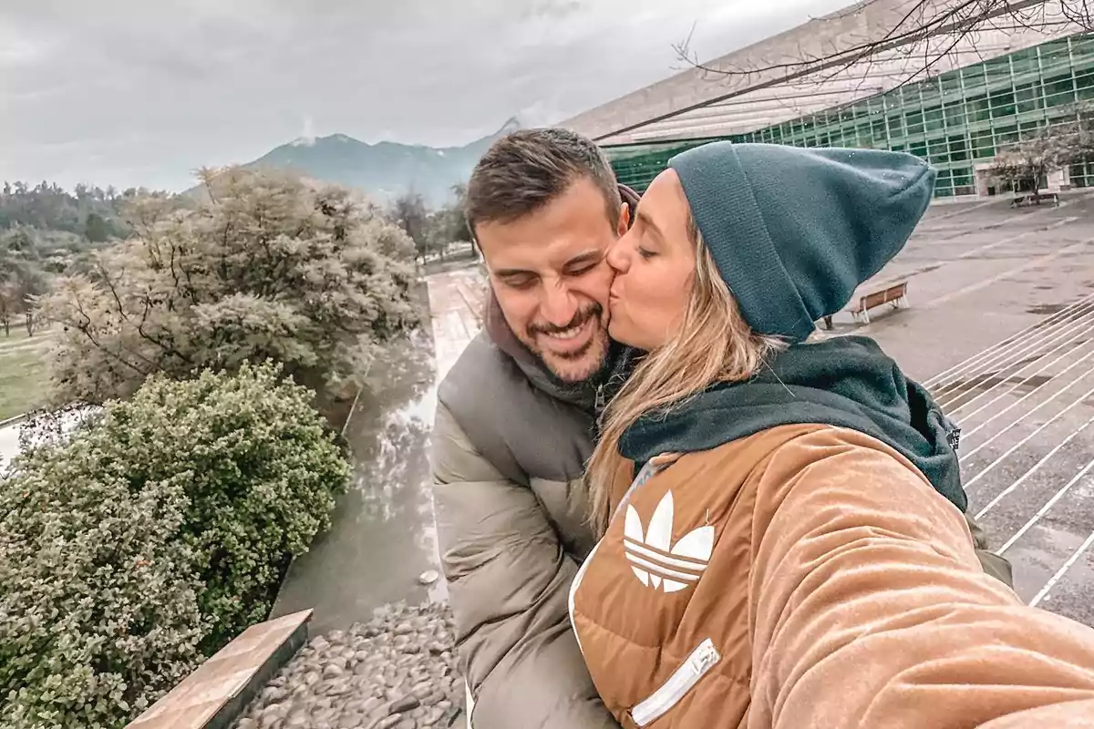 Una pareja se toma una selfie mientras se abrazan y sonríen en un entorno al aire libre con árboles y un edificio de fondo.