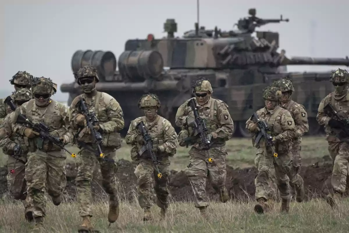 Soldados en uniforme de camuflaje corren en formación con un tanque en el fondo.