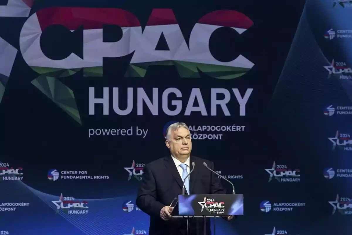 Un hombre de traje está de pie en un podio frente a un fondo que dice "CPAC Hungary" con gráficos y logotipos relacionados.