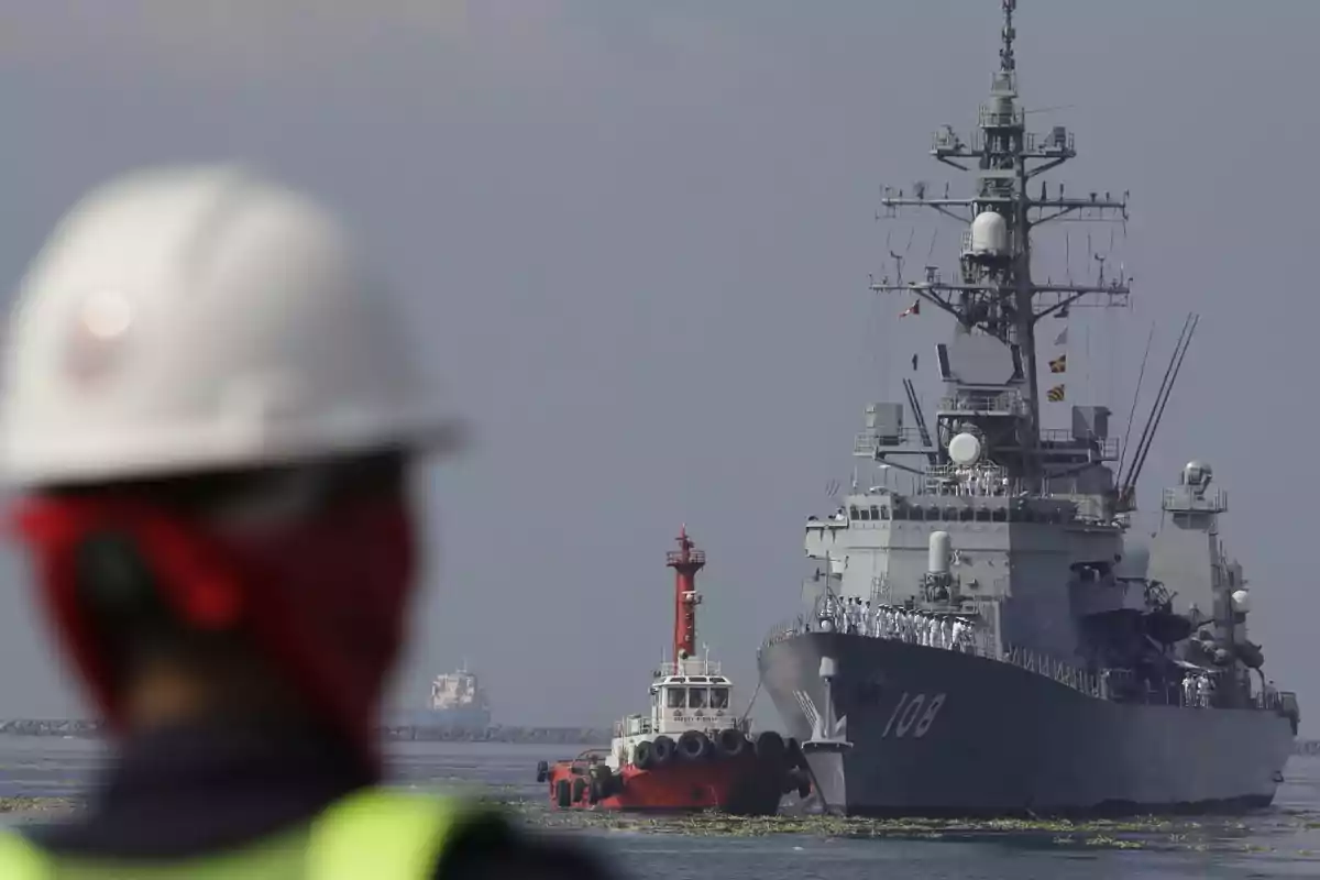 Un trabajador con casco observa un buque de guerra y un remolcador en el mar.