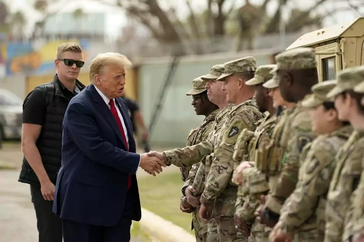 Un hombre de traje azul y corbata roja estrecha la mano de un soldado mientras otros militares observan, y un hombre con gafas de sol está de pie cerca.