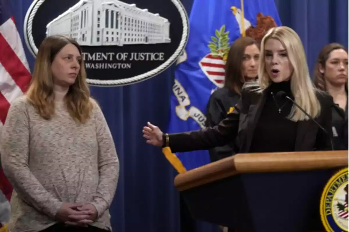 A woman speaking at a podium during a press conference at the United States Department of Justice, with several people standing behind her.