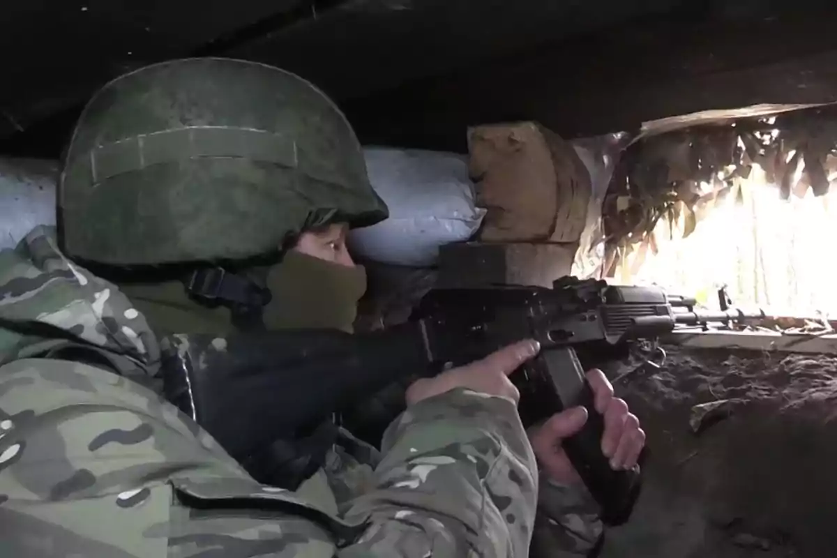 A soldier in camouflage uniform and helmet holds a rifle while observing through an opening in a trench.