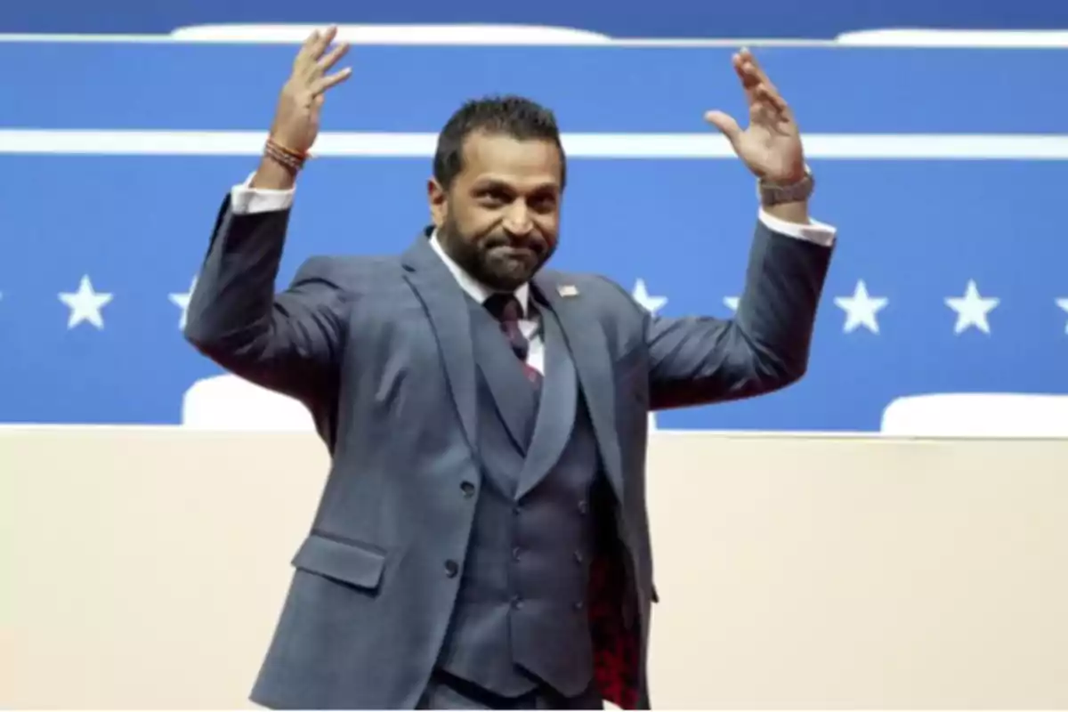 Un hombre con traje levanta las manos en un escenario con fondo azul y estrellas blancas.