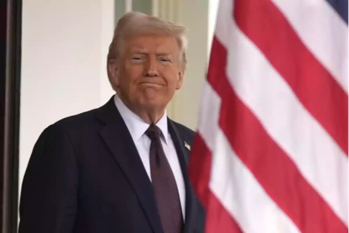 Un hombre de traje oscuro y corbata está de pie junto a una bandera de Estados Unidos.