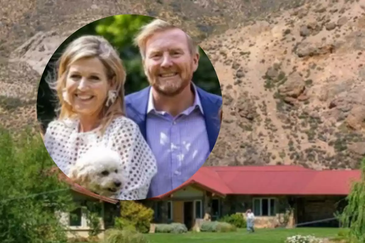 Una pareja sonriente con un perro en brazos frente a un paisaje montañoso y una casa con techo rojo.