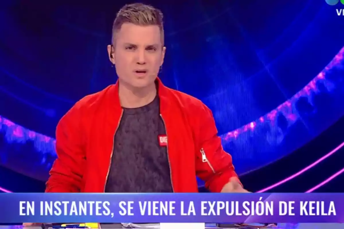 Un presentador de televisión con chaqueta roja habla en un programa con un anuncio en pantalla que dice "En instantes, se viene la expulsión de Keila".