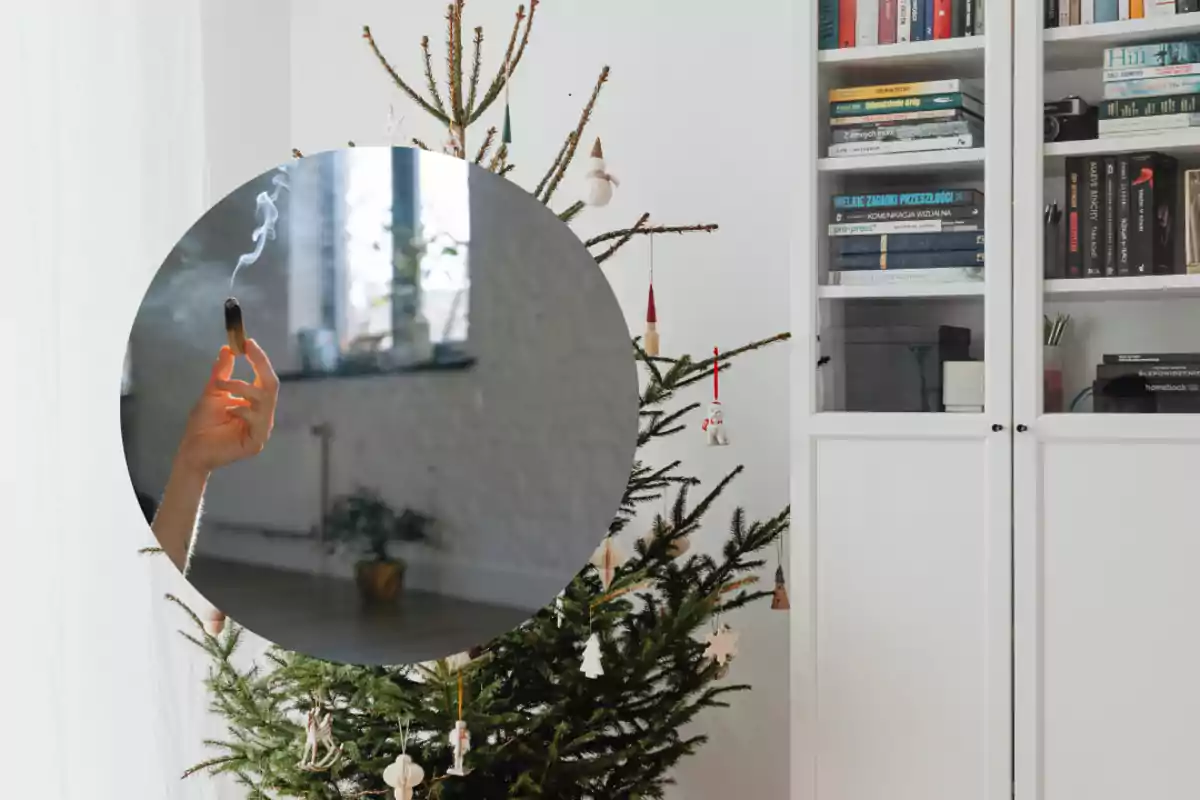 Un árbol de Navidad decorado junto a un mueble con libros y una mano sosteniendo un incienso encendido.