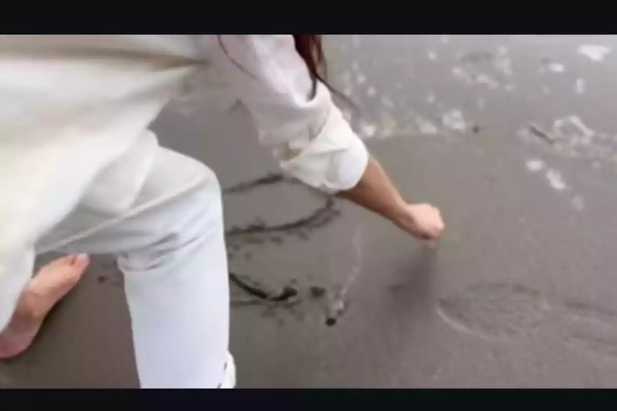 Persona dibujando en la arena húmeda de la playa con el dedo mientras está descalza y vestida de blanco.