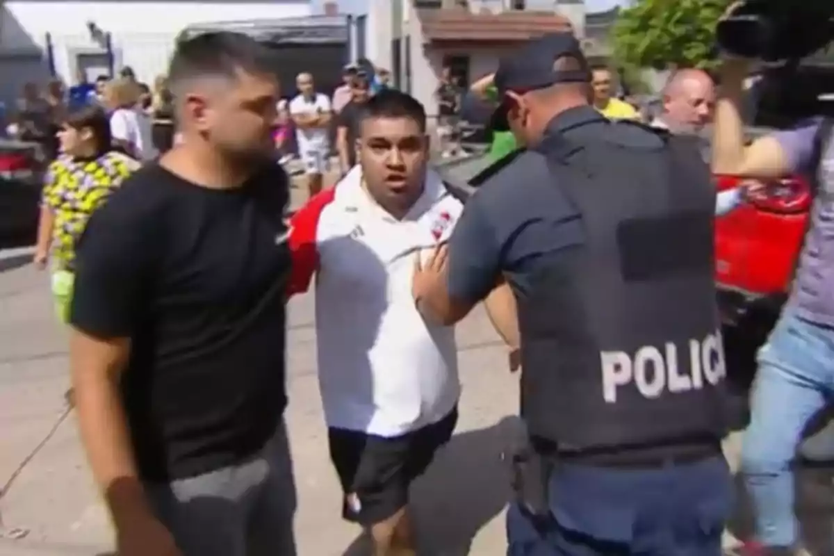 Un hombre con camiseta blanca es escoltado por un policía mientras varias personas observan en el fondo, en la casa del policia que mató a un colectivero.