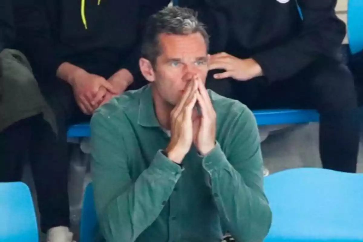 Un hombre con camisa verde está sentado en unas gradas azules con las manos juntas frente a su rostro en actitud pensativa.