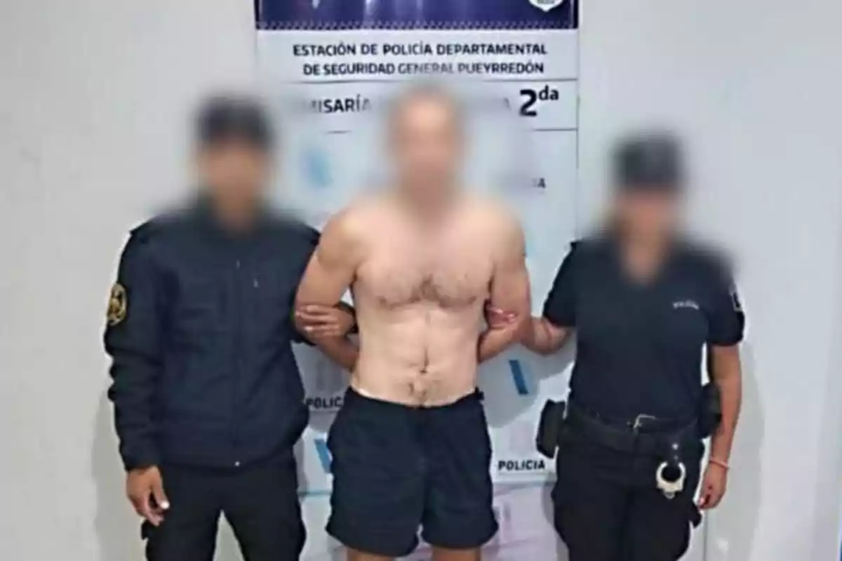 Tres personas de pie, con dos oficiales de policía flanqueando a un hombre sin camisa, frente a un cartel de una estación de policía.