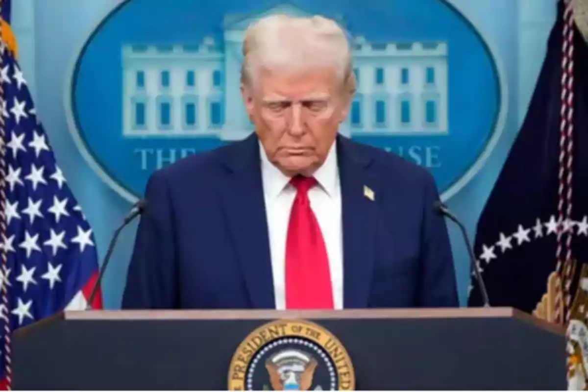 Un hombre de traje azul y corbata roja está de pie frente a un podio con el sello presidencial de Estados Unidos, con banderas a los lados y un fondo que muestra un edificio gubernamental.