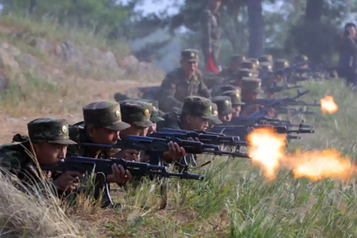 Soldados en uniforme camuflado disparando rifles en posición de cuerpo a tierra durante un ejercicio militar en un campo abierto.