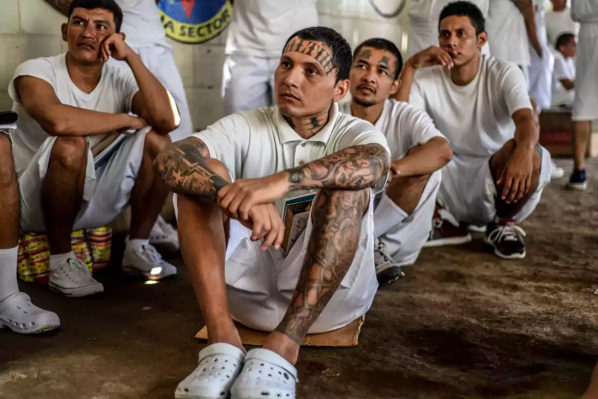 Un grupo de hombres con ropa blanca y tatuajes visibles están sentados en el suelo en una habitación.