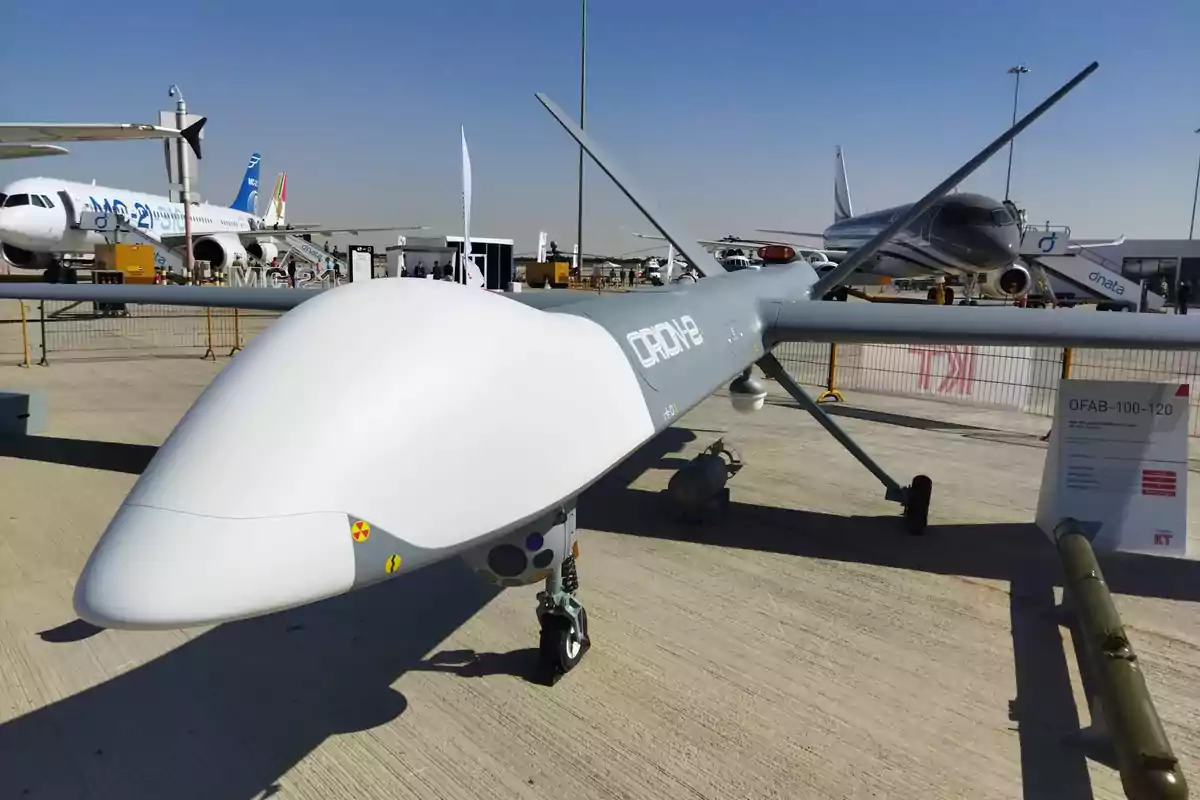 Un dron grande exhibido en una feria de aviación con aviones comerciales en el fondo.