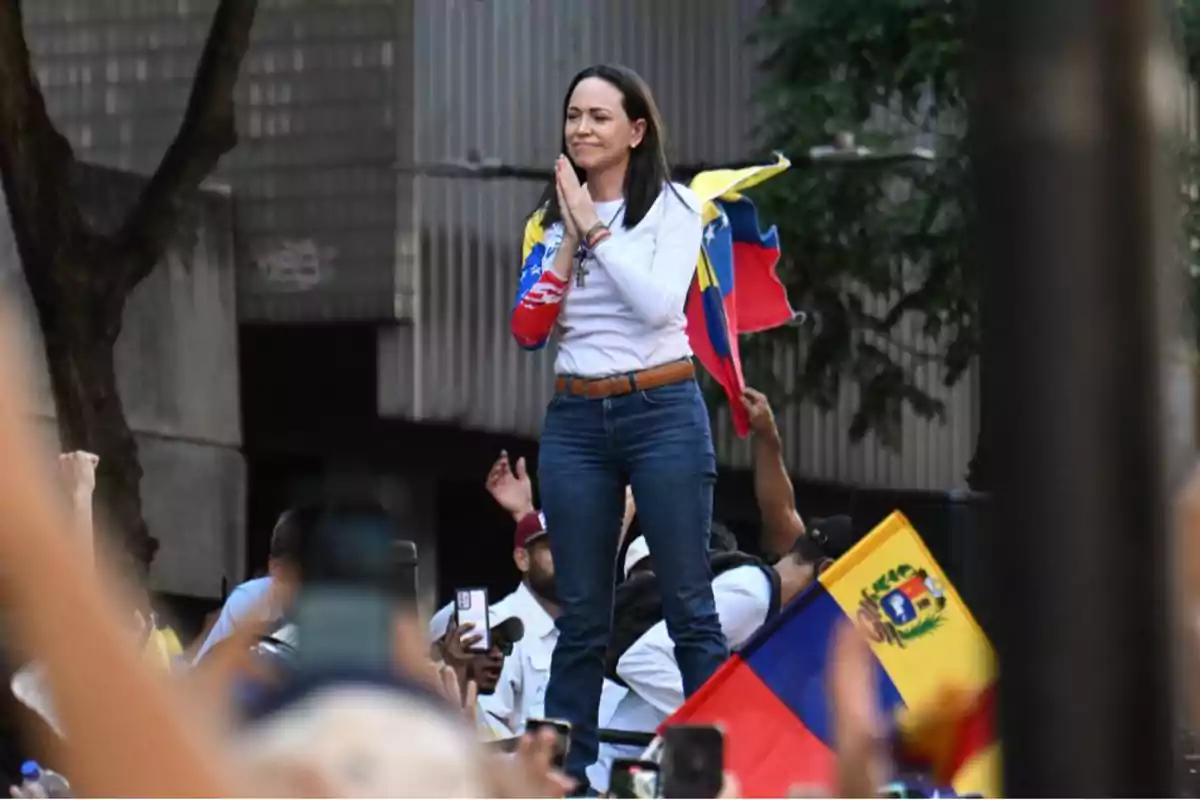 Una mujer con una bandera de Venezuela sobre los hombros se encuentra de pie en una manifestación rodeada de personas que sostienen banderas venezolanas.