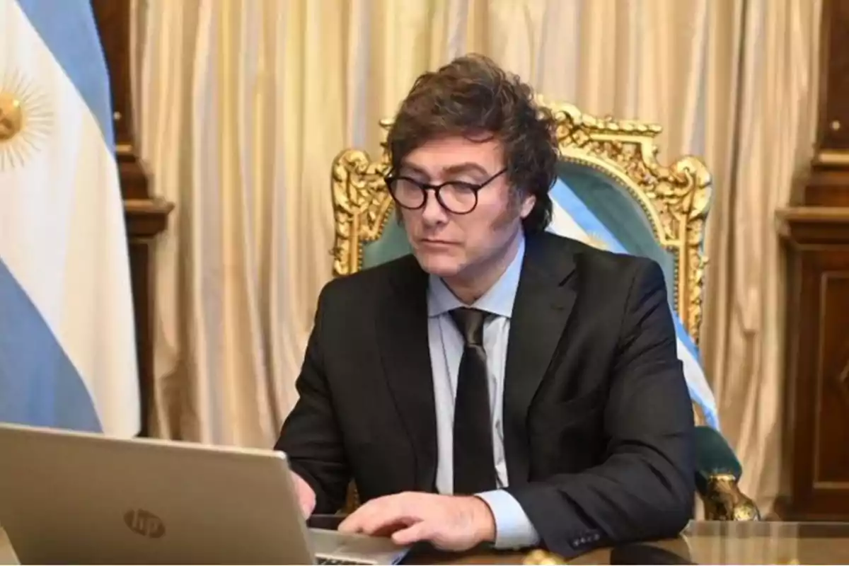 Un hombre con gafas y traje oscuro trabaja en una computadora portátil en una oficina elegante con una bandera argentina de fondo.