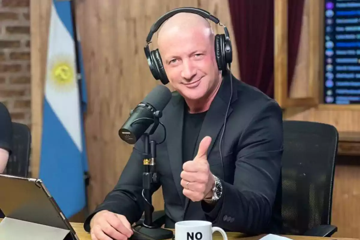 Un hombre calvo con auriculares y un micrófono, vestido de negro, hace un gesto de aprobación con el pulgar levantado mientras está sentado en un estudio de grabación con una bandera de Argentina en el fondo.