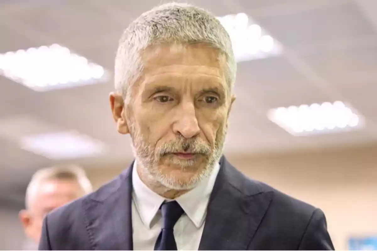 Un hombre de cabello canoso y barba vestido con traje oscuro y corbata en un entorno interior iluminado.