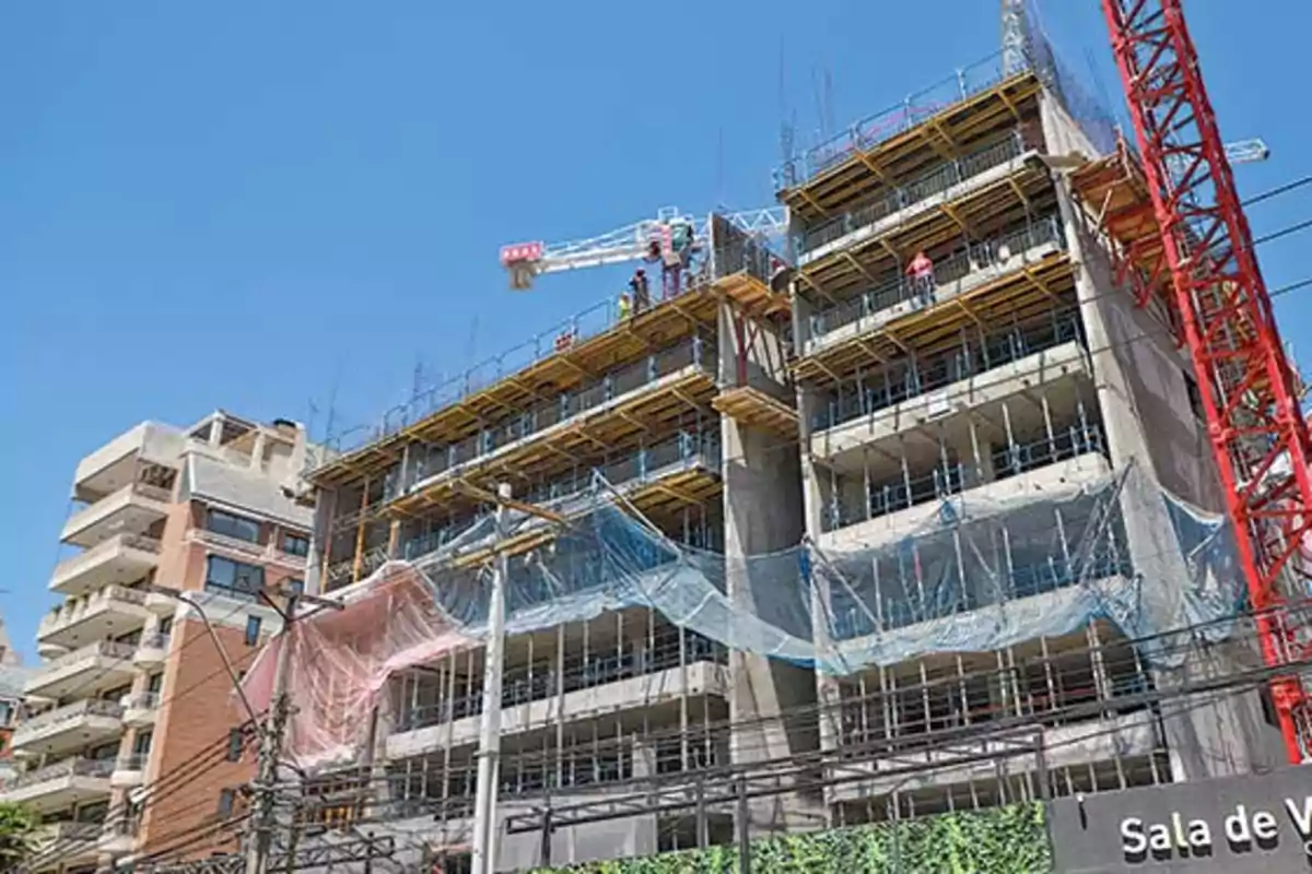 Edificio en construcción con andamios y redes de seguridad, junto a una grúa roja y cielo despejado.