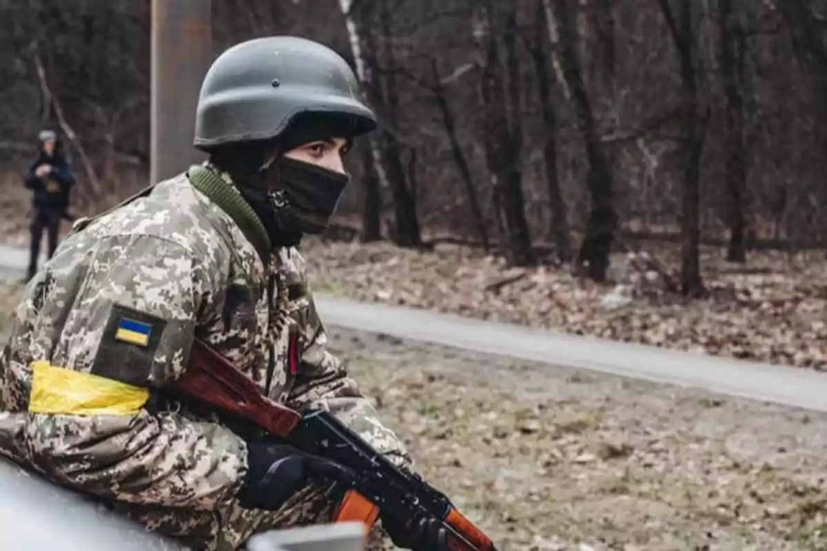 Un soldado con uniforme de camuflaje y casco, portando un rifle, se encuentra en un entorno boscoso.