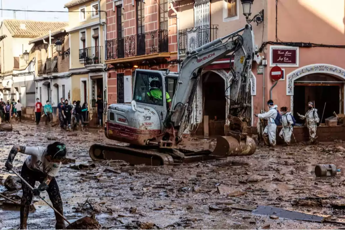 Una excavadora y personas con equipo de protección limpian una calle llena de barro y escombros después de una inundación en un área urbana.