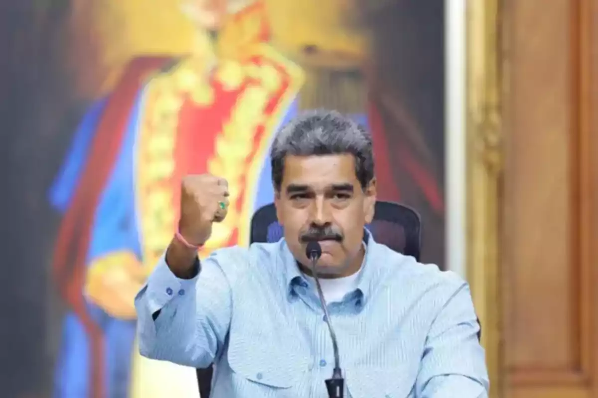 Un hombre con camisa azul levanta el puño mientras habla frente a un micrófono con un retrato colorido en el fondo.