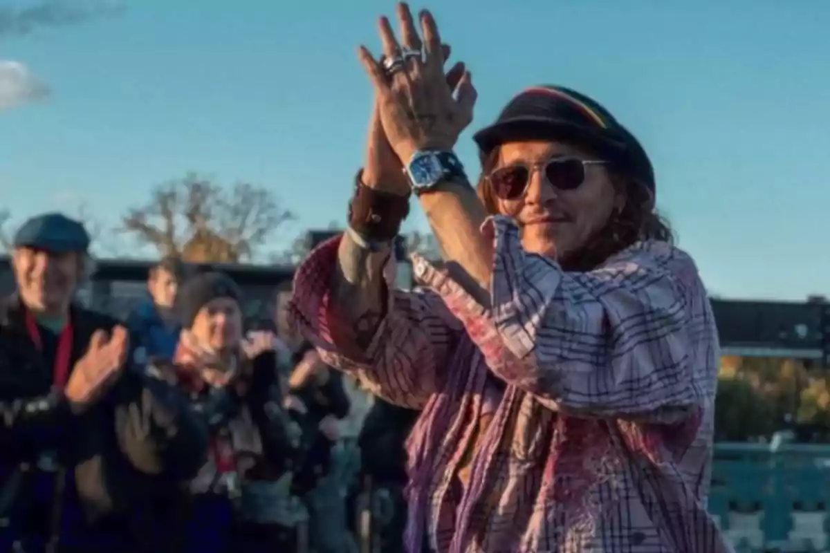 Una persona con sombrero y gafas de sol aplaudiendo al aire libre mientras otras personas aplauden en el fondo.