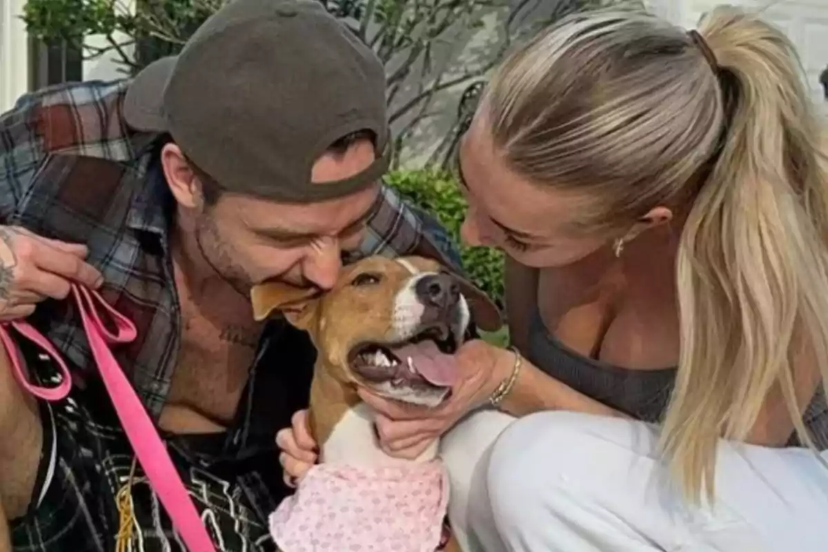 Una pareja sonriente acaricia a un perro que lleva un pañuelo rosa.