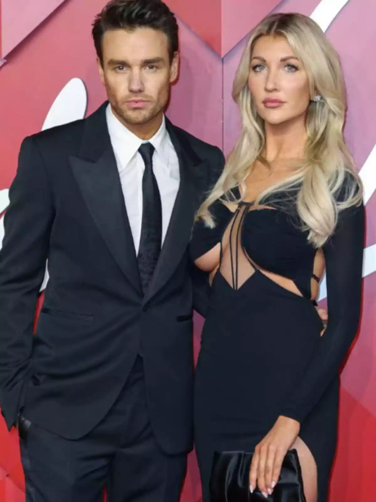 Dos personas posando en un evento formal con fondo rojo, el hombre lleva un traje negro y la mujer un vestido negro elegante.