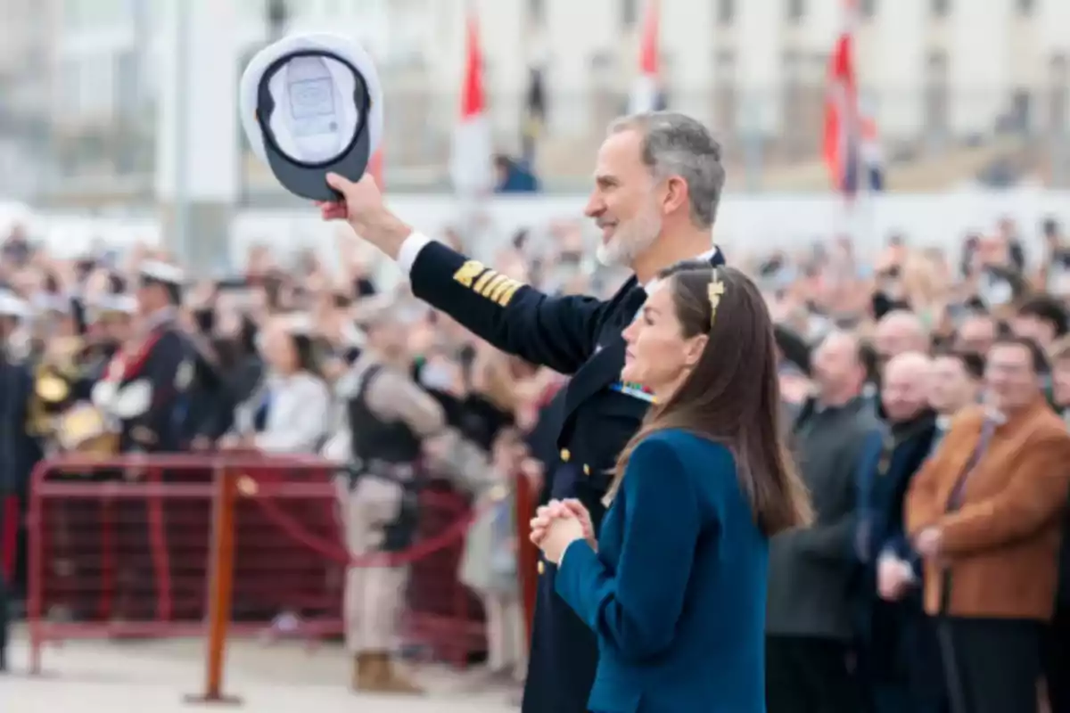 Un hombre con uniforme militar levanta su gorra mientras una mujer a su lado observa, rodeados de una multitud en un evento al aire libre.