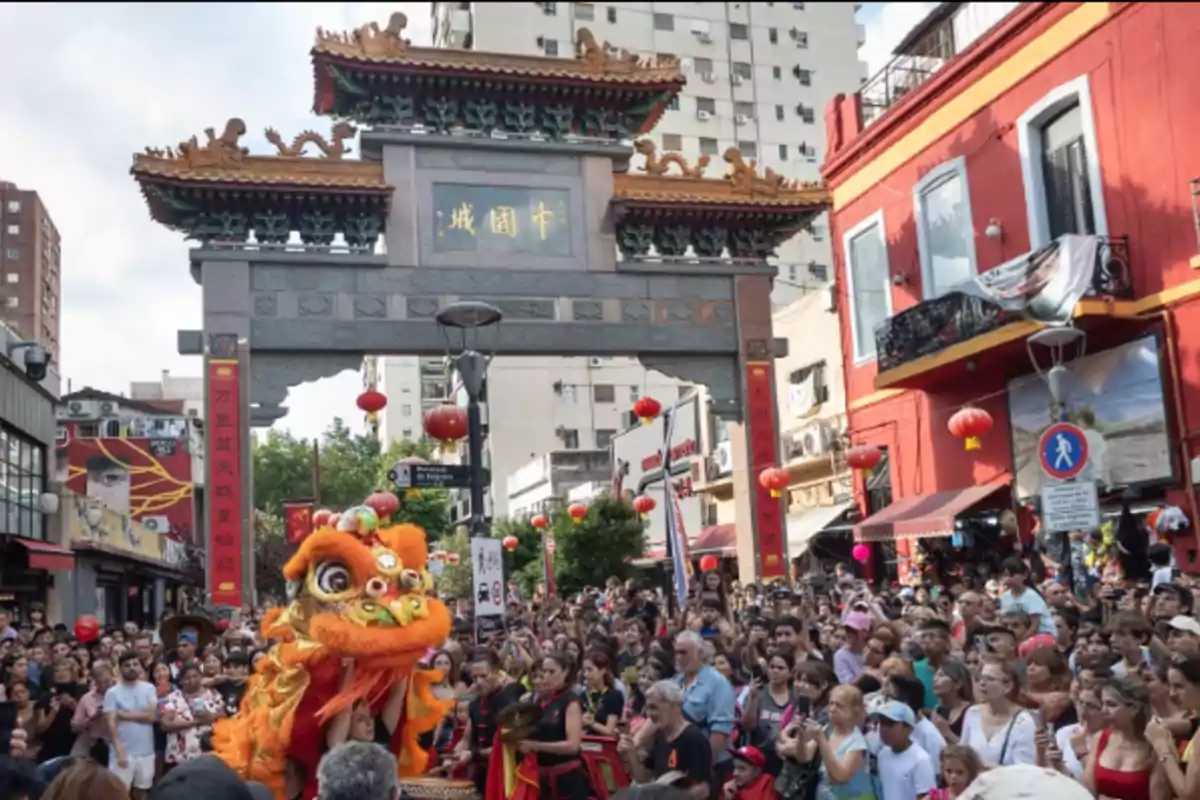 Una multitud celebra el Año Nuevo Chino en un barrio con decoraciones tradicionales y un arco chino, mientras un grupo realiza una danza del león.