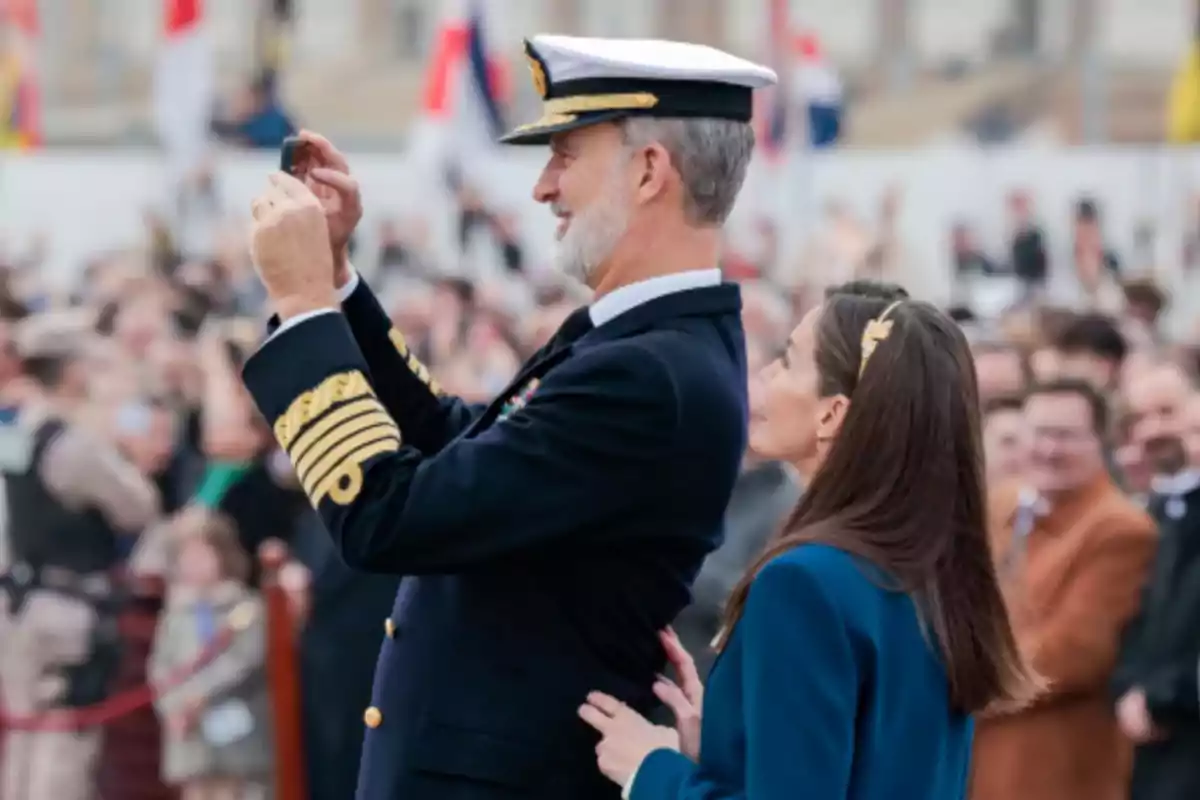 Un hombre con uniforme naval toma una foto con su teléfono mientras una mujer a su lado lo observa en un evento al aire libre con una multitud de fondo.