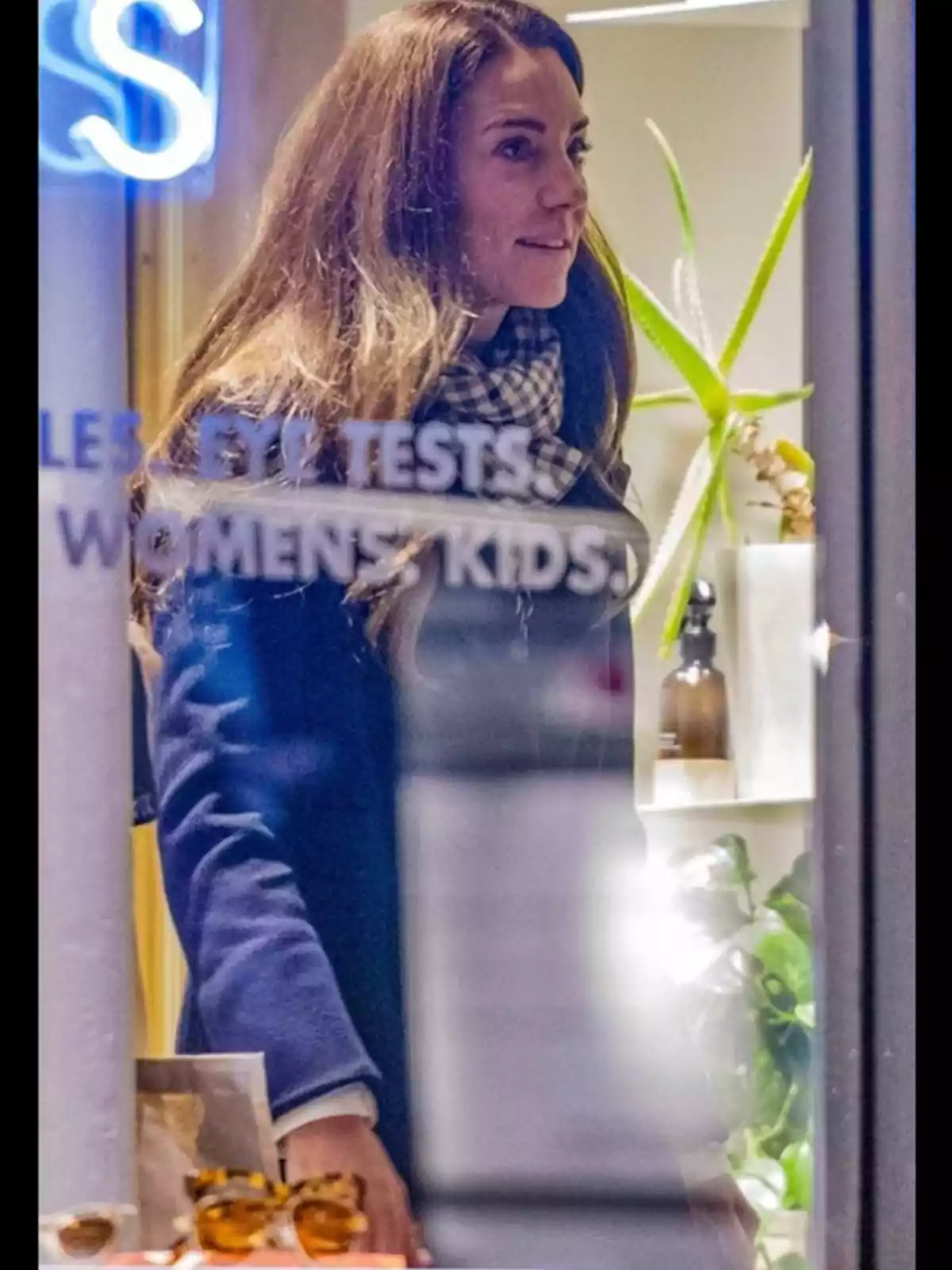 Una mujer con abrigo azul y bufanda a cuadros está dentro de una tienda, observada a través de una ventana con texto parcialmente visible y plantas en el fondo.