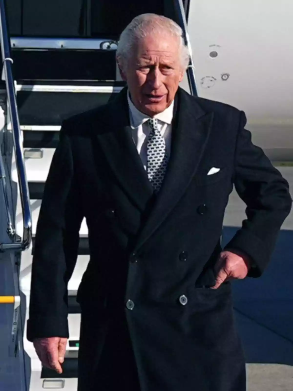 Un hombre mayor con abrigo oscuro y corbata de pie frente a unas escaleras de avión.