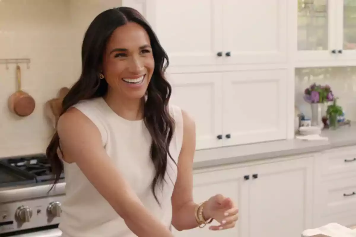 Una mujer sonriente en una cocina moderna con gabinetes blancos y utensilios de cocina en el fondo.