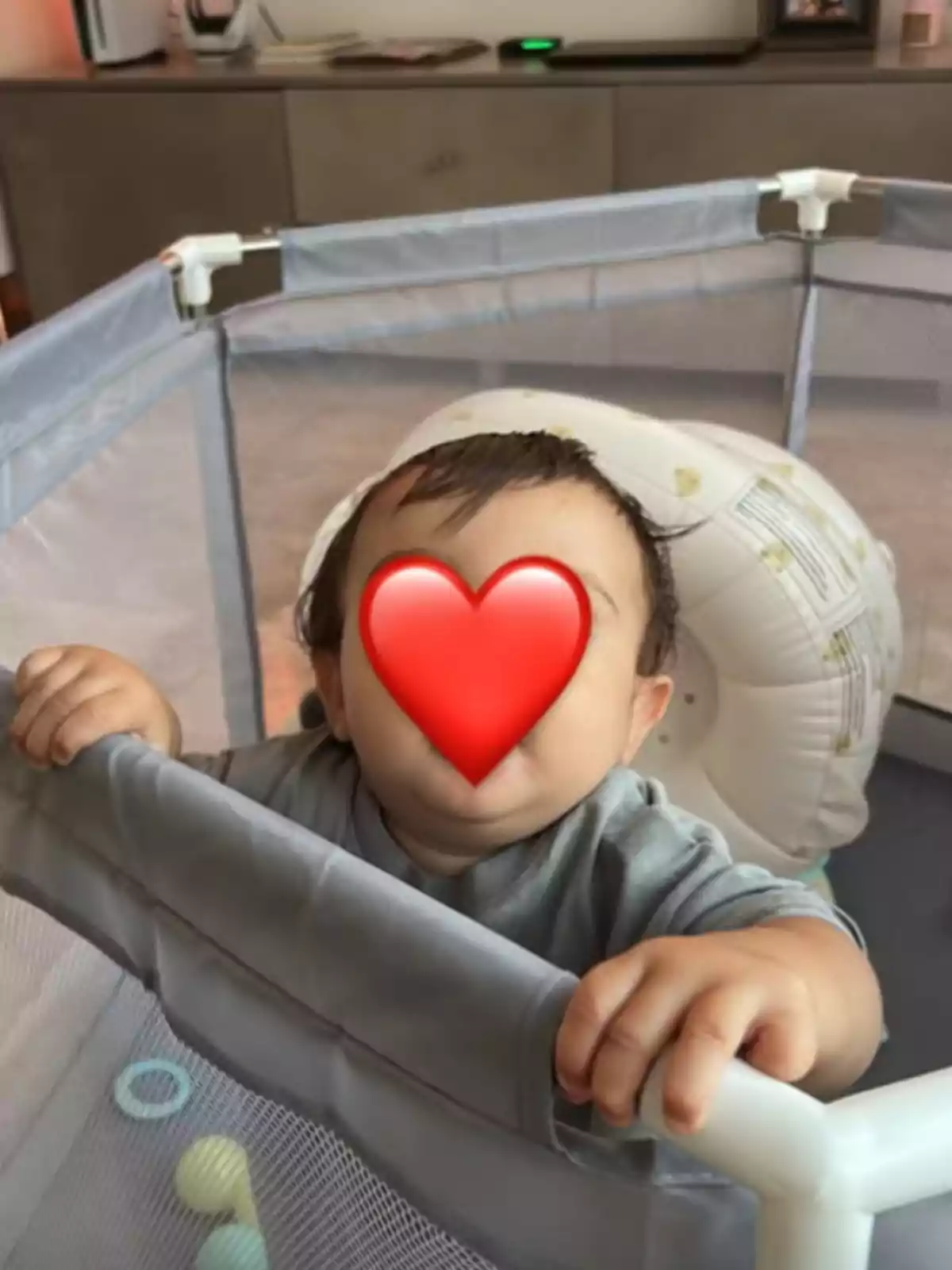 Un bebé en un corralito con un emoji de corazón cubriendo su rostro.