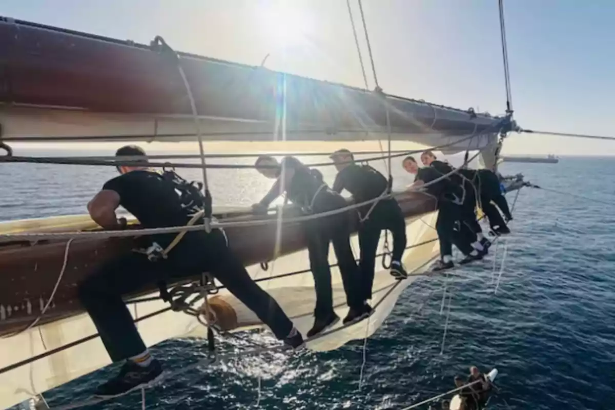 Un grupo de personas trabaja en el aparejo de un barco sobre el mar, con el sol brillando en el horizonte.