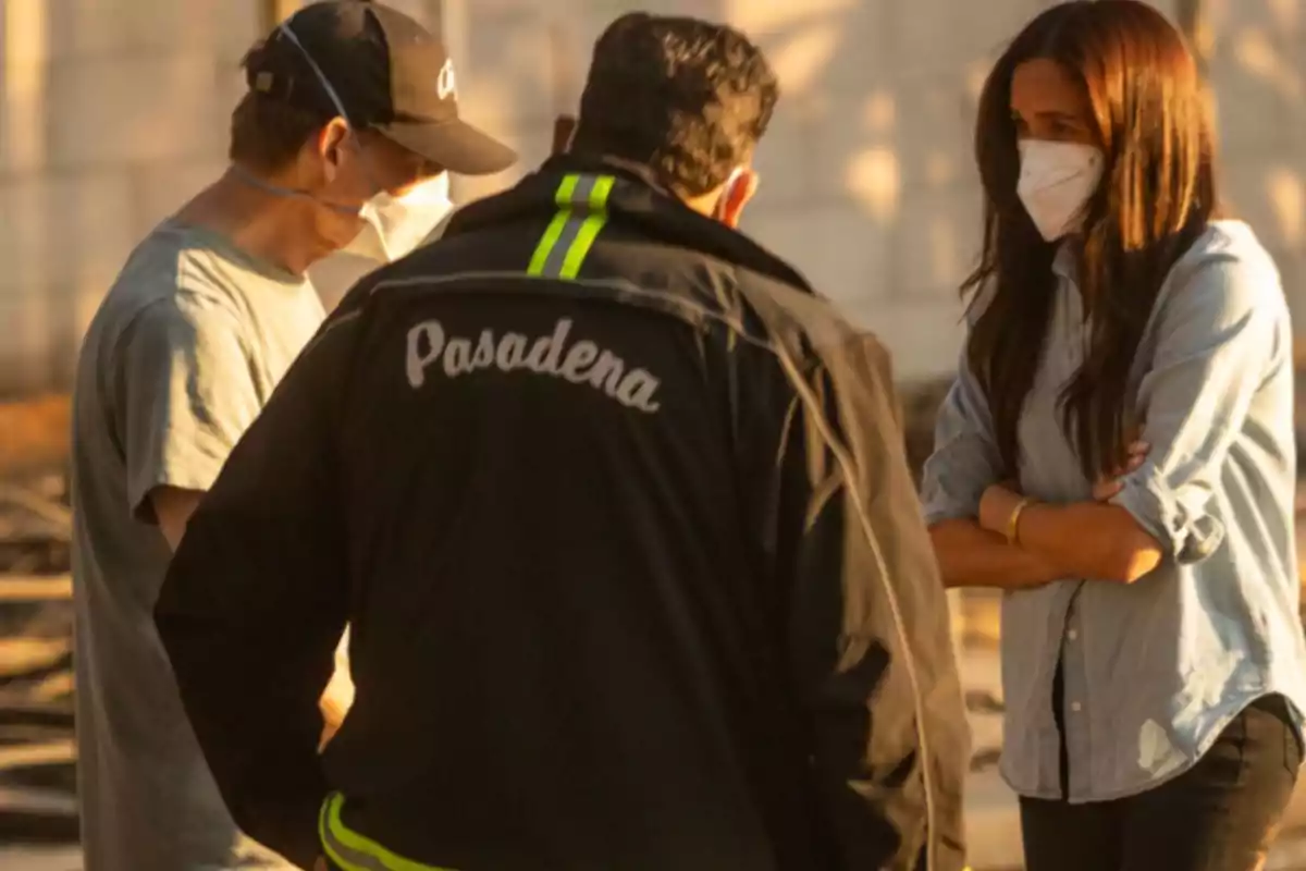 Tres personas con mascarillas conversan al aire libre, una de ellas lleva una chaqueta con la palabra 