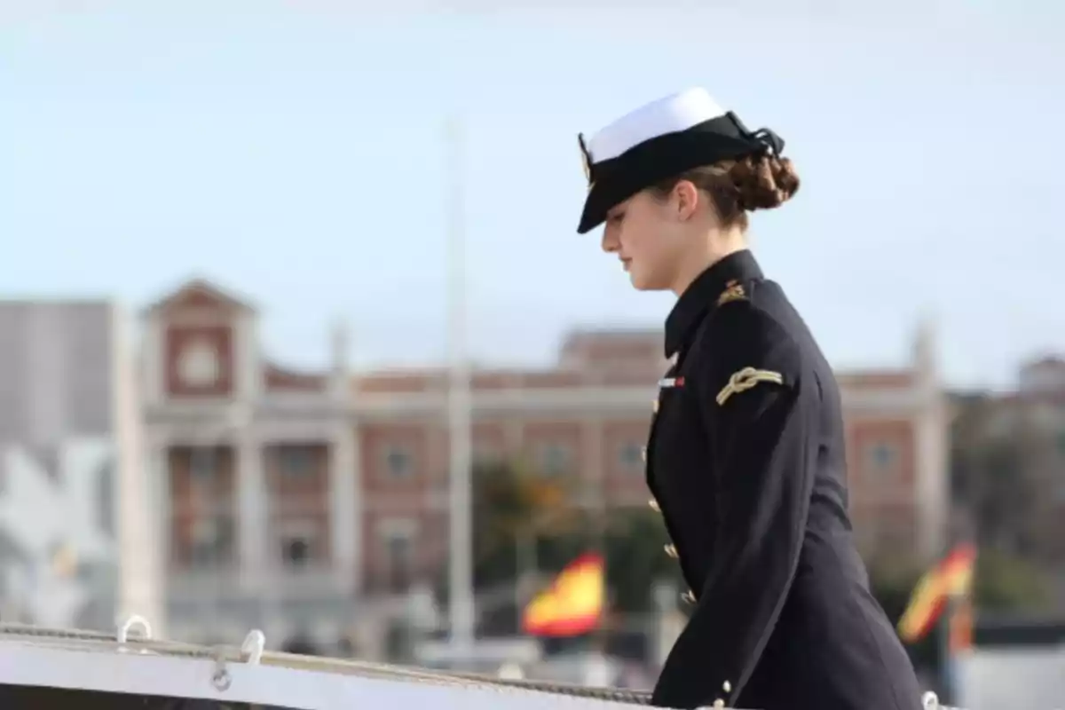 La princesa Leonor con uniforme naval camina en un entorno urbano con banderas de España al fondo.