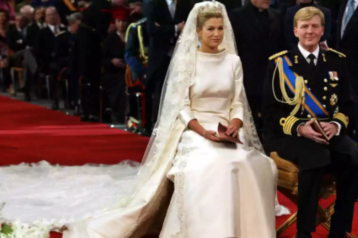 Una pareja vestida de manera formal, con la mujer usando un vestido de novia y el hombre un uniforme militar, sentados en un evento ceremonial.