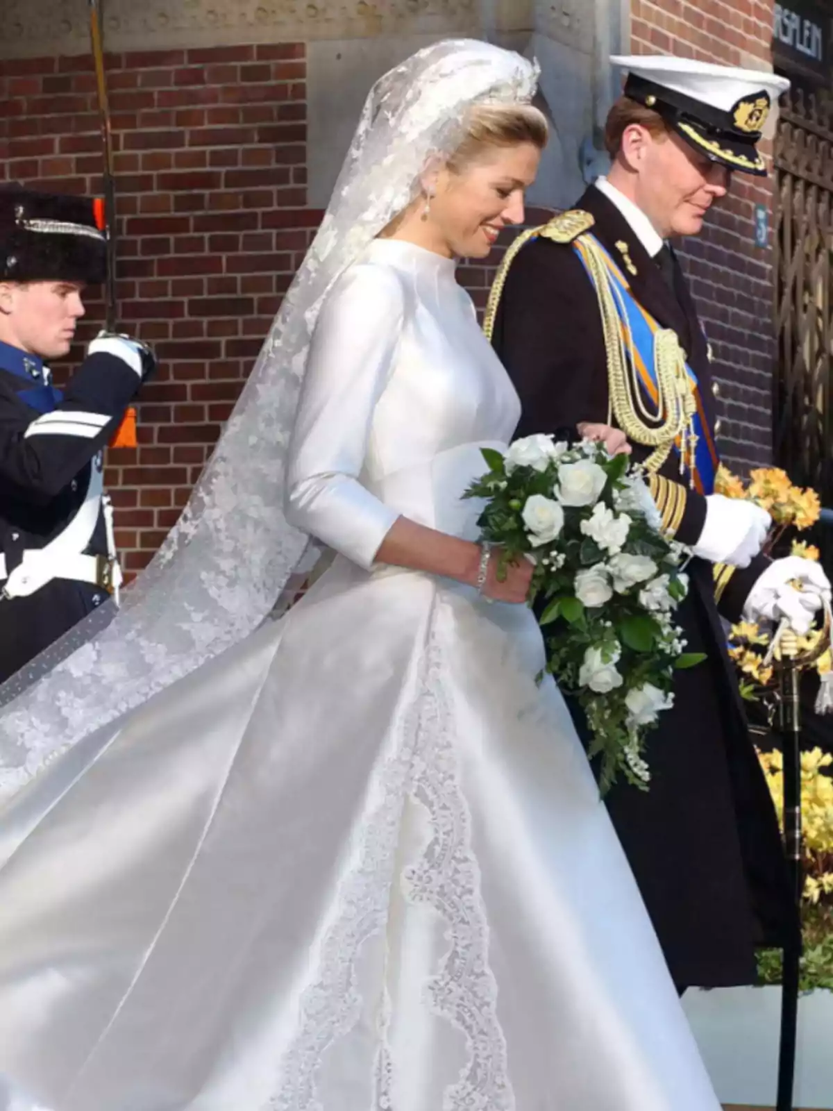 Una pareja vestida de novios camina juntos, ella lleva un vestido blanco con encaje y un ramo de flores, mientras él viste un uniforme militar con gorra.