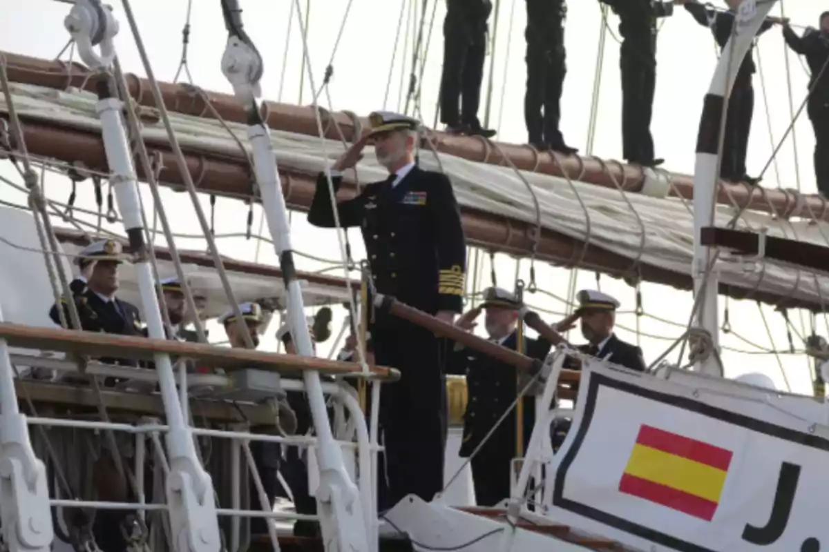 Un grupo de oficiales navales saluda en la cubierta de un barco con una bandera de España visible.