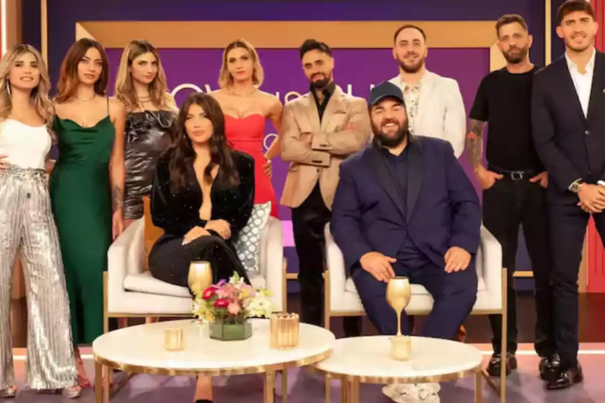 A group of people posing on a television set, some standing and others sitting, with tables decorated with flowers and glasses in front.